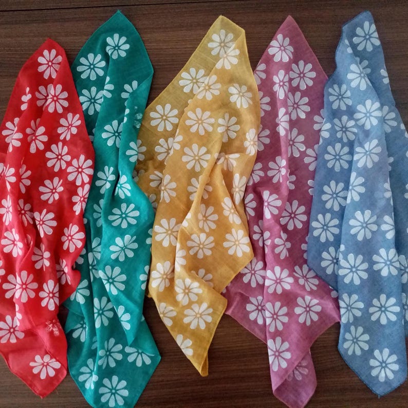 May include: Five colorful bandanas with a white daisy pattern. The bandanas are in shades of red, teal, yellow, pink, and blue. The fabric is lightweight and appears to be cotton or a similar material. The bandanas are folded and arranged in a row.