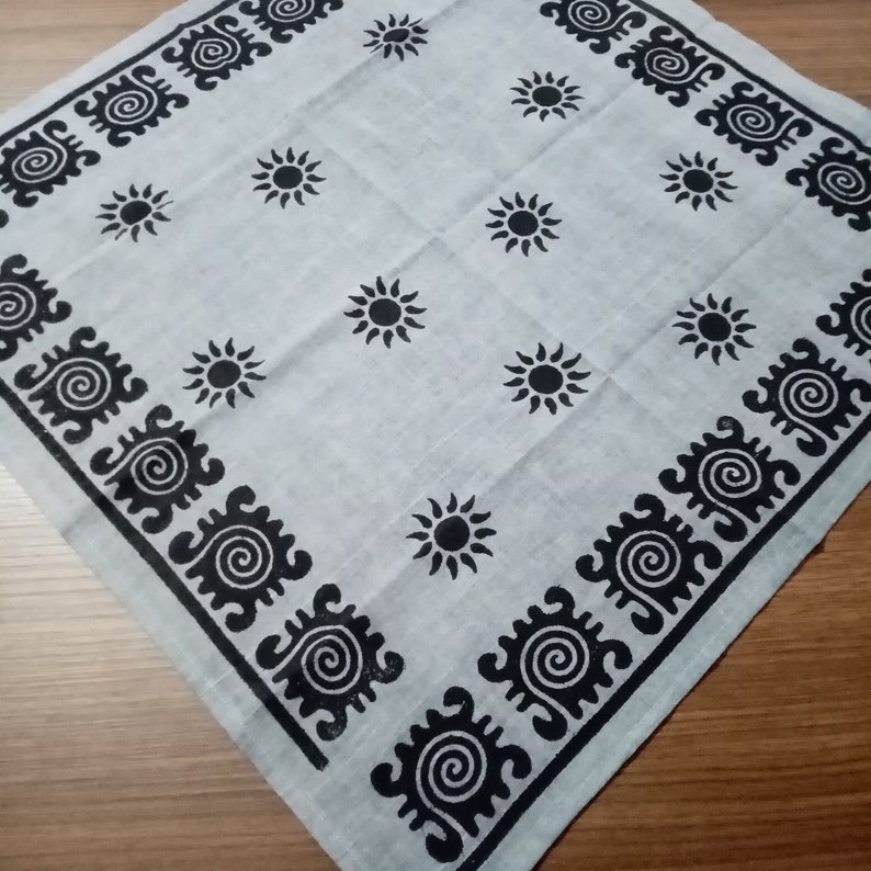 May include: A square, light gray bandana with a repeating pattern of black suns and a black border with a spiral design. The bandana is made of a soft, lightweight fabric and is folded on a wooden surface.