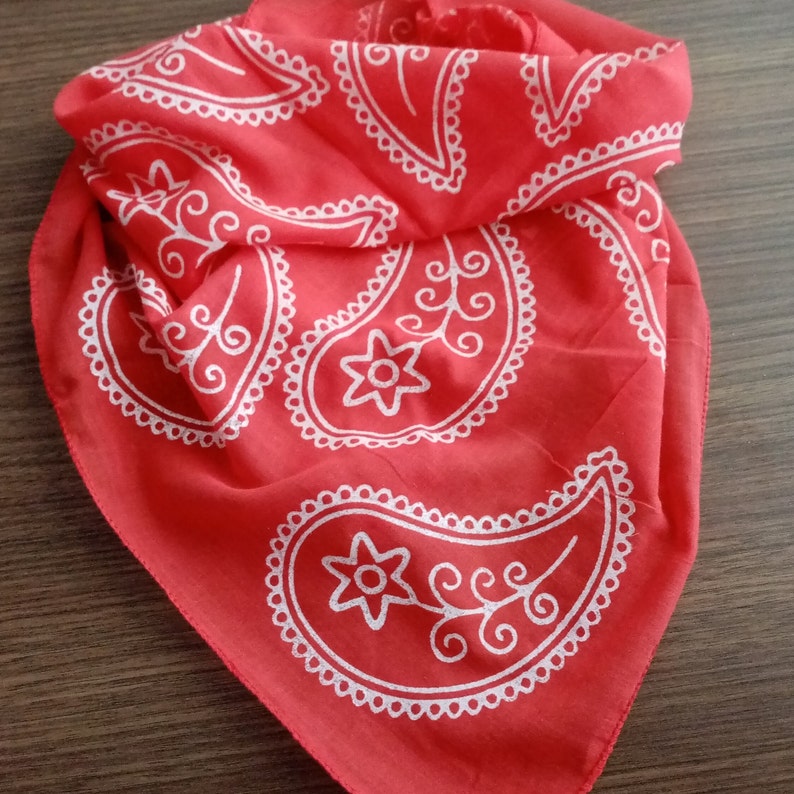 May include: Red bandana with a white paisley pattern. The bandana is folded and has a fringed edge.