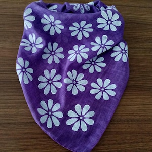 May include: A purple bandana with a repeating pattern of white daisy-like flowers. The fabric is folded, showing the front and part of the back. The bandana is made of a soft material.