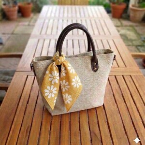 May include: A beige tote bag with brown leather handles and accents. A yellow scarf with white daisy patterns is tied to the handle. The bag is sitting on a wooden table.