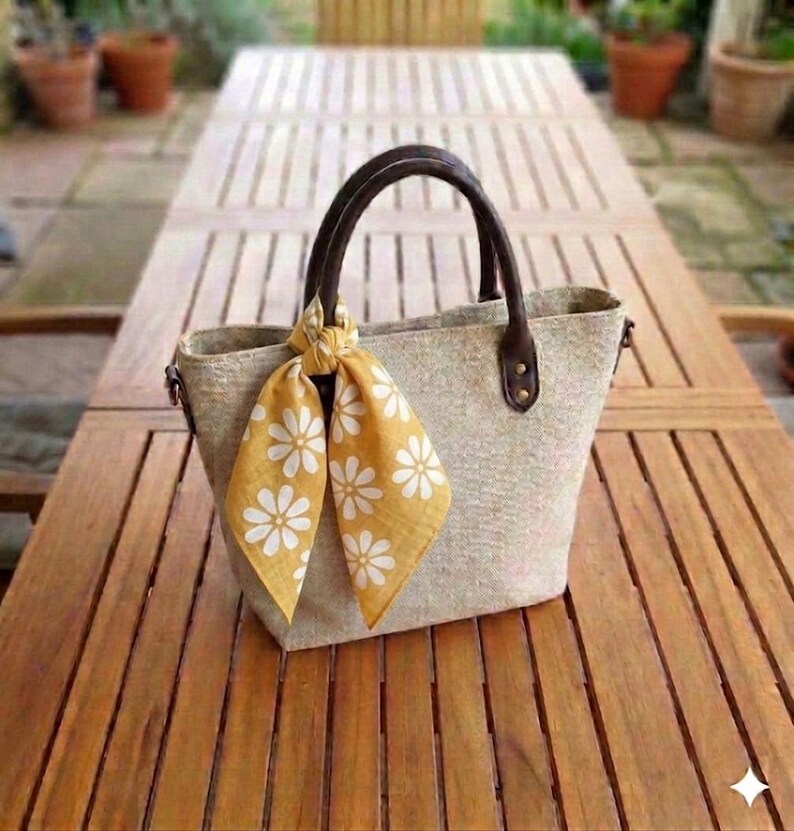 May include: A beige tote bag with brown leather handles and accents. A yellow scarf with white daisy patterns is tied to the handle. The bag is sitting on a wooden table.