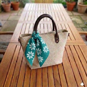 May include: A beige tote bag with brown leather handles and a teal scarf adorned with white daisy patterns. The bag is made of a woven material and sits on a wooden table, suggesting a casual, summery theme.