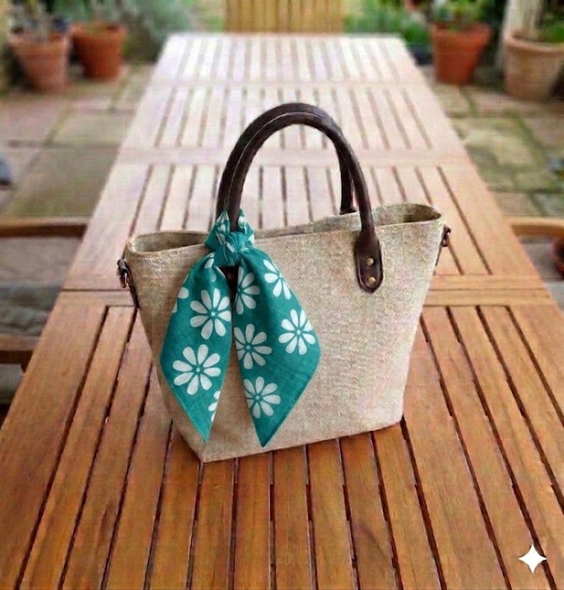 May include: A beige tote bag with brown leather handles and a teal scarf adorned with white daisy patterns. The bag is made of a woven material and sits on a wooden table, suggesting a casual, summery theme.