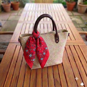 May include: A structured tote bag made of a light-colored, textured woven material with two dark brown handles. A vibrant red scarf featuring a detailed floral pattern in shades of blue, green, and white is tied around one of the bag's handles.