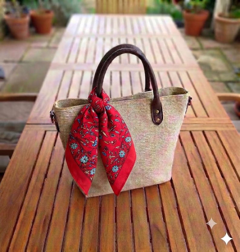 May include: A structured tote bag made of a light-colored, textured woven material with two dark brown handles. A vibrant red scarf featuring a detailed floral pattern in shades of blue, green, and white is tied around one of the bag's handles.
