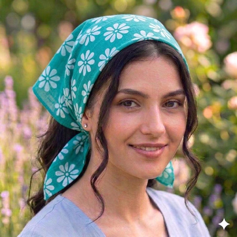 May include: A teal headscarf with a white daisy pattern. The head covering is tied at the back and worn on the head. The background is a blurred garden with pink and purple flowers.