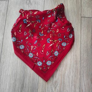 May include: A square scarf in a deep red hue featuring a repeating pattern of stylized flowers and vines in shades of light blue, white, and dark red. The design includes a distinct border of small blue and white blossoms along the edges.
