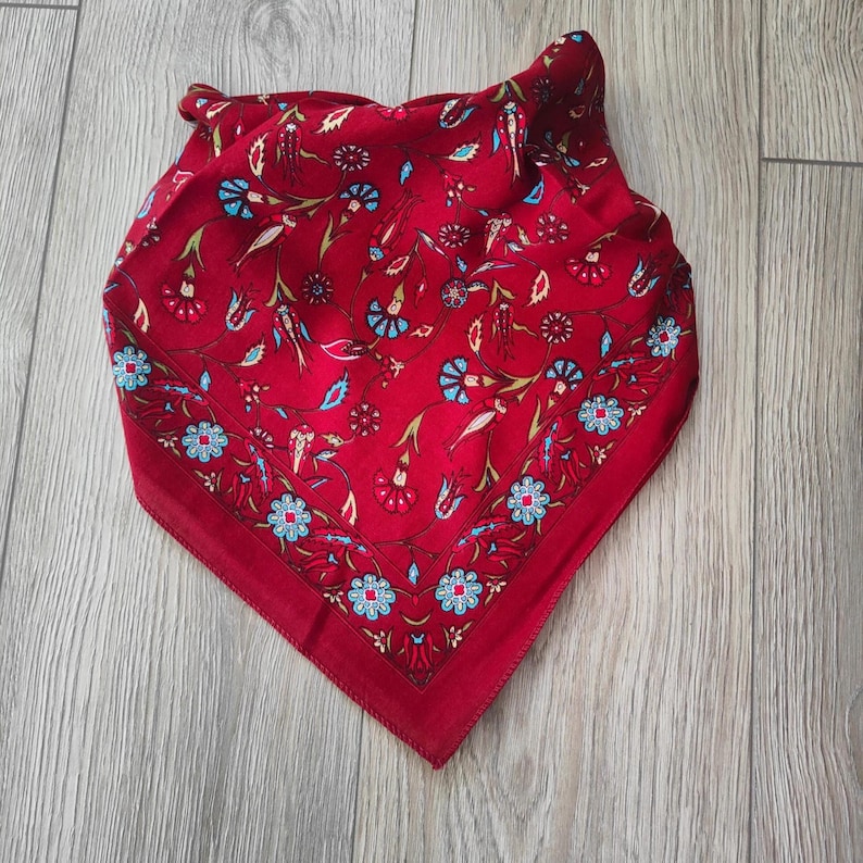 May include: A square scarf in a deep red hue featuring a repeating pattern of stylized flowers and vines in shades of light blue, white, and dark red. The design includes a distinct border of small blue and white blossoms along the edges.