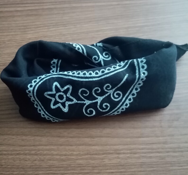 May include: A black bandana with a white paisley design. The bandana is folded and lying on a wooden surface.