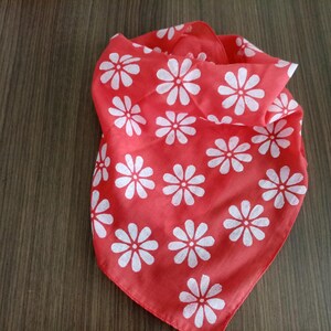 May include: A red bandana with a white daisy pattern. The square scarf is folded and displayed on a wooden surface. The floral design is evenly spaced across the fabric.