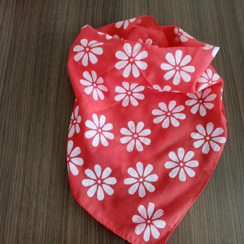 May include: A red bandana with a white daisy pattern. The square scarf is folded and displayed on a wooden surface. The floral design is evenly spaced across the fabric.