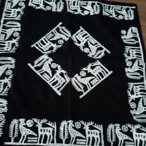 May include: Black square textile with white embroidered folk art designs. The border features stylized deer and trees. The center has four diamond-shaped designs with deer and bird motifs. A decorative piece.