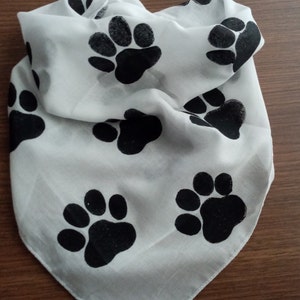 Pode incluir: Bandana branca com um padr&atilde;o repetido de pegadas pretas. O tecido est&aacute; dobrado, mostrando a textura e o design. Adequada para animais de estima&ccedil;&atilde;o ou como acess&oacute;rio de moda.
