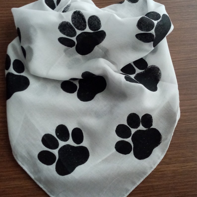 Pode incluir: Bandana branca com um padr&atilde;o repetido de pegadas pretas. O tecido est&aacute; dobrado, mostrando a textura e o design. Adequada para animais de estima&ccedil;&atilde;o ou como acess&oacute;rio de moda.