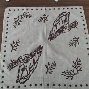 K&ouml;nnte beinhalten: Eine quadratische, beige Stoffserviette mit einem dunkelbraunen Druckmuster aus zwei Pferden und mehreren Blattzweigen. Der Rand der Serviette ist mit einem sich wiederholenden dunkelbraunen Punktmuster entlang aller vier Kanten des Textils verziert.