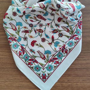 May include: A white square scarf featuring a repeating floral pattern with red tulips, blue carnations, and green stems. The design includes a decorative border of blue and red flowers that frames the central motif of scattered botanical elements.