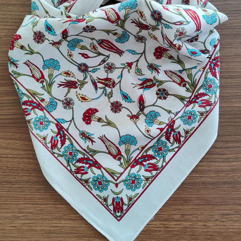 May include: A white square scarf featuring a repeating floral pattern with red tulips, blue carnations, and green stems. The design includes a decorative border of blue and red flowers that frames the central motif of scattered botanical elements.
