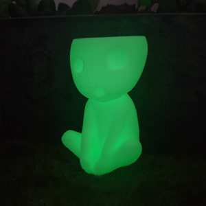 May include: A glowing green figurine of a small, humanoid figure sitting with its legs crossed. The figurine is made of a smooth, white material and has a simple, minimalist design.