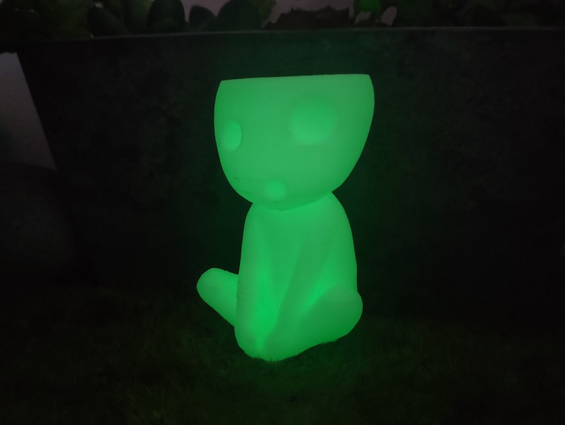 May include: A glowing green figurine of a small, humanoid figure sitting with its legs crossed. The figurine is made of a smooth, white material and has a simple, minimalist design.