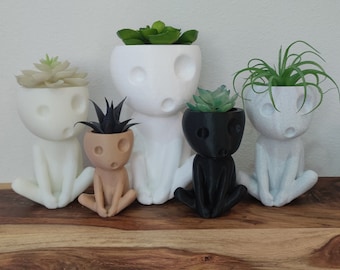 May include: Five white, black, and brown ceramic planters shaped like human figures with different plants growing inside. The planters are sitting on a wooden surface.