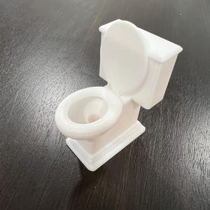 May include: A small, white toilet model with the lid up. The miniature toilet is made of a solid material and sits on a dark surface. The toilet bowl and tank are clearly defined, showcasing a detailed design.