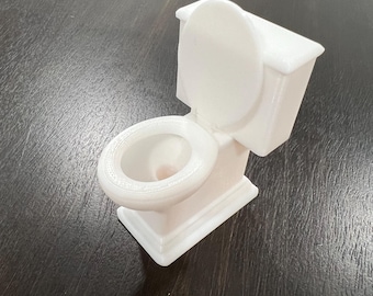 May include: A small, white toilet model with the lid up. The miniature toilet is made of a solid material and sits on a dark surface. The toilet bowl and tank are clearly defined, showcasing a detailed design.