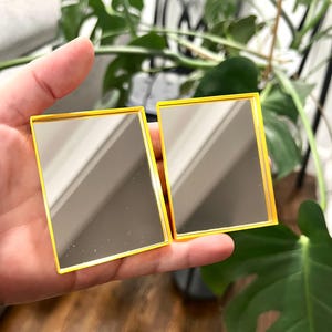 May include: Two small, rectangular mirrors with yellow frames. The mirrors are held in a hand, reflecting the surrounding environment. The mirrors are a simple, minimalist design.