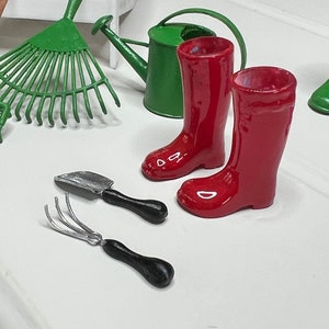 May include: A collection of miniature garden tools, including a green rake, a green watering can, a small trowel, a hand fork, and two glossy red rain boots. The tools are arranged on a white surface.