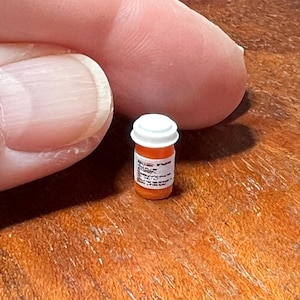 May include: Miniature orange pill bottle with a white cap. The bottle has a label with illegible text. The bottle is small, likely a dollhouse accessory or a novelty item. The bottle is on a wooden surface.