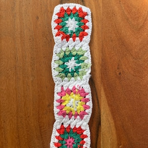 Könnte beinhalten: Handgefertigter Häkel-Lesezeichen mit vier Granny Squares. Jeder Quadrat hat einen weißen Rand und ein zentrales Blumendesign in Rot, Grün, Rosa und Gelb. Das Lesezeichen wird auf einer Holzoberfläche präsentiert.