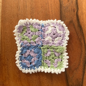Könnte beinhalten: Ein quadratischer, handgefertigter Häkeluntersetzer mit weißem, gewelltem Rand. Der Untersetzer ist in vier Abschnitte unterteilt, jeder mit einem anderen Farbschema: Blau, Grün, Weiß und Lavendel. Der Untersetzer liegt auf einer Holzoberfläche.
