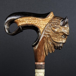 May include: A wooden walking stick with a carved buffalo head handle. The handle is made of dark wood with a light brown buffalo head carved into it. The stick is made of light brown wood with a dark brown band around the top.