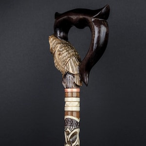 Op de afbeelding: Een houten wandelstok met een gesneden uil ontwerp en een donkerbruine handgreep. De handgreep is hartvormig en heeft een koperen band.