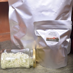 May include: Two silver resealable bags of freeze-dried shredded cabbage. The bags are labeled "Freeze Dried Organically Grown Shredded Cabbage" and "Lucas Organic Farms". A glass jar filled with the freeze-dried cabbage is in the foreground.