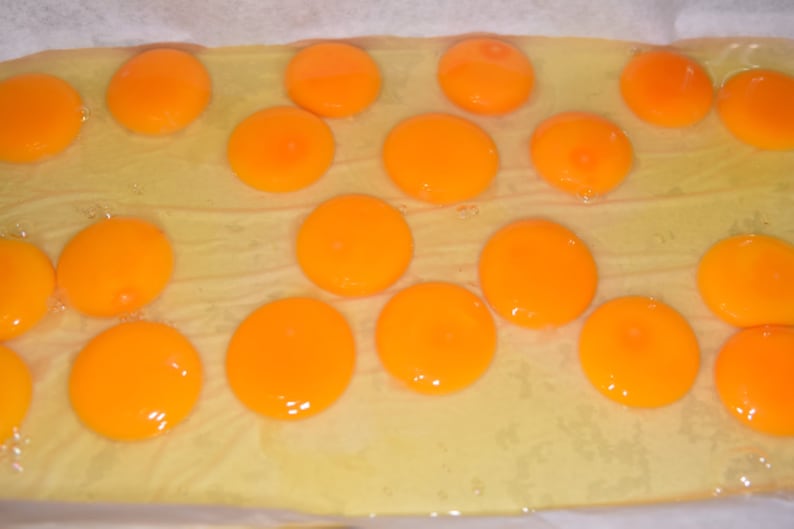 May include: A close-up of a baking sheet with 14 bright orange egg yolks. The yolks are arranged in a pattern on a white parchment paper-lined baking sheet.