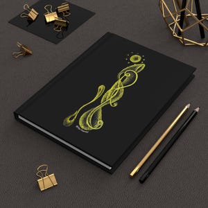 May include: A black notebook with a gold design of a stylized swirl with a circle at the top. The notebook is on a gray surface with two pencils and a gold paperclip.