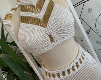 May include: A white macrame two-piece outfit with gold accents. The top features a high neck and geometric design with gold bars. The skirt has a fringed hemline and gold detailing at the waist. The outfit is displayed on a mannequin.