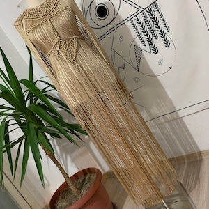 Könnte beinhalten: Ein langes, beigefarbenes Häkelkleid mit Fransen. Das Kleid wird auf einer Schaufensterpuppe vor einer weißen Wand mit geometrischem Muster präsentiert.