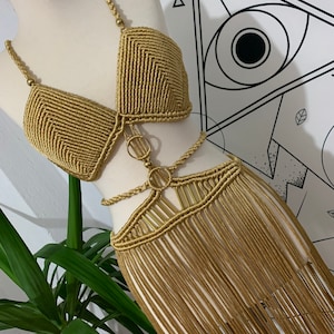 以下が含まれることがあります： マネキンに展示された、ゴールドのマクラメビキニトップとスカートのセット。 ビキニトップはトライアングルカップとビーズストラップが特徴です。 スカートはフリンジデザインです。 セットは織りコードで作られています。