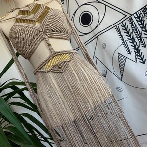 Op de afbeelding: Een beige macramé top en rok set met gouden accenten. De top is halter stijl en de rok heeft lange franjes.