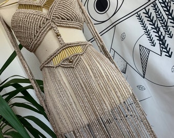 Könnte beinhalten: Ein beigefarbenes Macrame-Oberteil und Rock-Set mit goldenen Akzenten. Das Oberteil ist im Halterstil gehalten und der Rock hat lange Fransen.