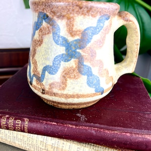 May include: A ceramic mug with a beige base, decorated with brown and blue abstract ribbon patterns. The mug has a curved handle. It is set against a backdrop of vintage books and greenery, suggesting a rustic or vintage aesthetic.