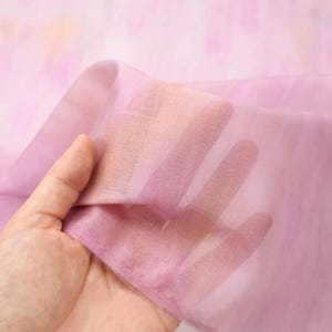 Op de afbeelding: Een doorschijnende stof in roze en paarse tinten wordt omhoog gehouden, waardoor de hand erachter zichtbaar wordt. De stof heeft een aquareleffect met gemengde kleuren. Het materiaal lijkt licht en delicaat, geschikt voor diverse knutselprojecten.