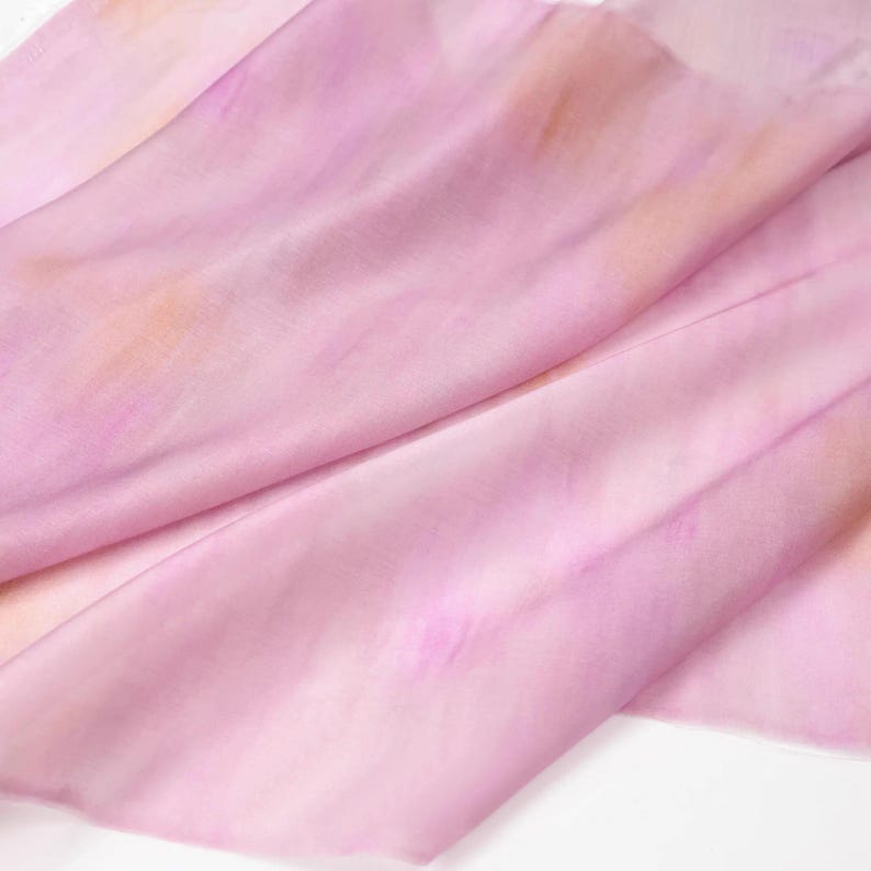 Op de afbeelding: Een zachte, vloeiende sjaal in roze en perziktinten. De stof lijkt zijde te zijn, met subtiele kleurvariaties die een aquareleffect cre&euml;ren. De sjaal is gedrapeerd, wat de delicate textuur en het potentieel voor elegante styling laat zien.