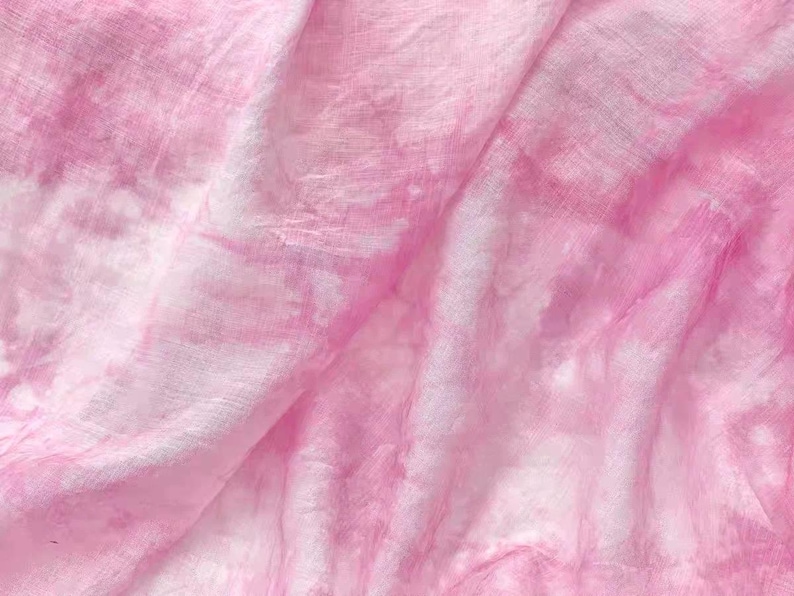 May include: A soft, pink fabric with a tie-dye pattern. The fabric is wrinkled and has a light, airy feel.