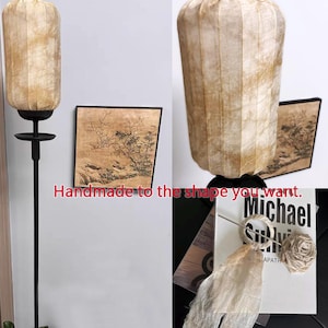May include: A beige lampshade with a pleated design hangs from a black metal stand. The lampshade is handmade and can be customized to different shapes. The image also shows a framed painting with a black frame and a beige background. The painting depicts a landscape with trees and birds.