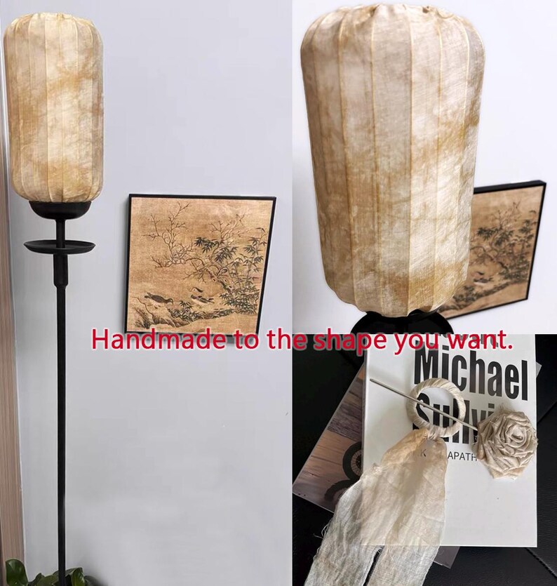 May include: A beige lampshade with a pleated design hangs from a black metal stand. The lampshade is handmade and can be customized to different shapes. The image also shows a framed painting with a black frame and a beige background. The painting depicts a landscape with trees and birds.