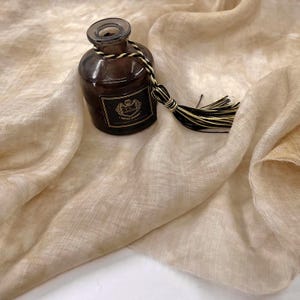 May include: A light brown linen scarf with a brown glass bottle with a black and white tassel.