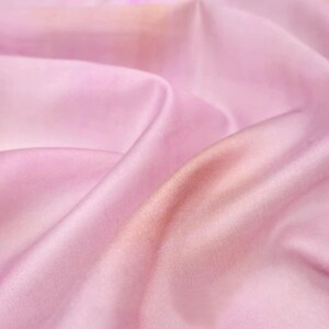 Op de afbeelding: Close-up van een vloeiende, lichtroze stof met subtiele kleurvariaties. De stof heeft zachte plooien en een gladde textuur, waardoor een zachte, etherische uitstraling ontstaat. De kleur gaat over van een zachtroze naar een warmere, perzikkleurige tint.